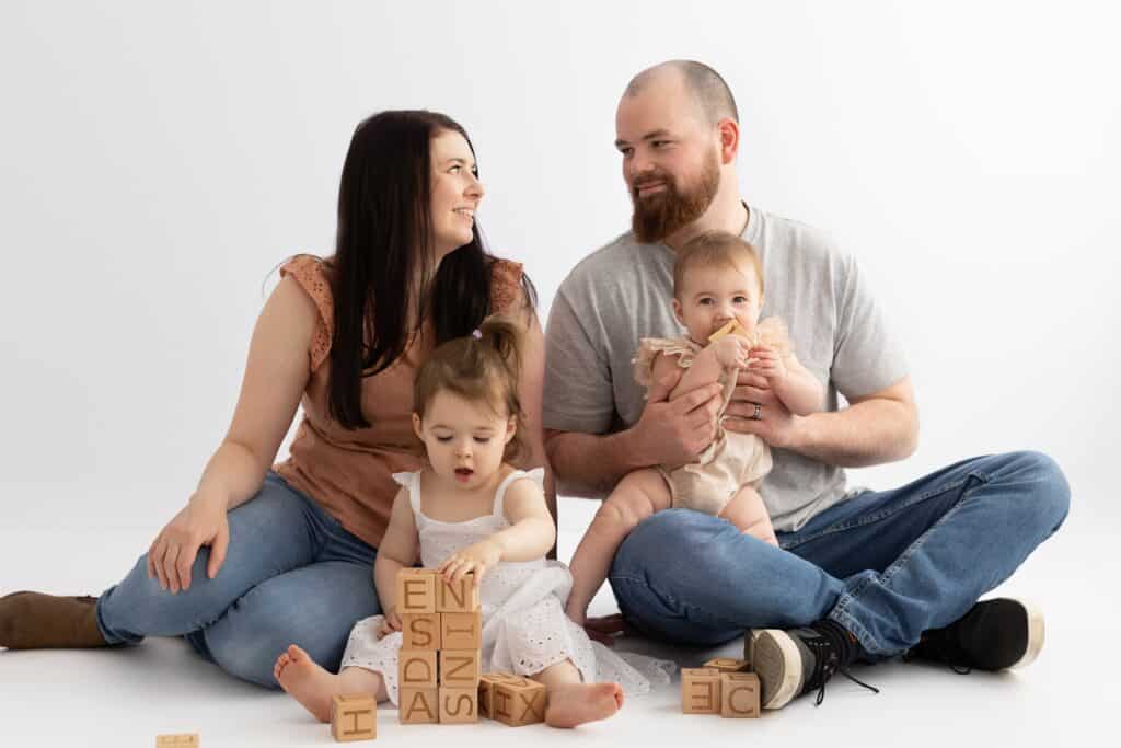 Two little girls play with wooden blocks with their parents looking at each other during family photography session in white studio in Pakenham Victoria