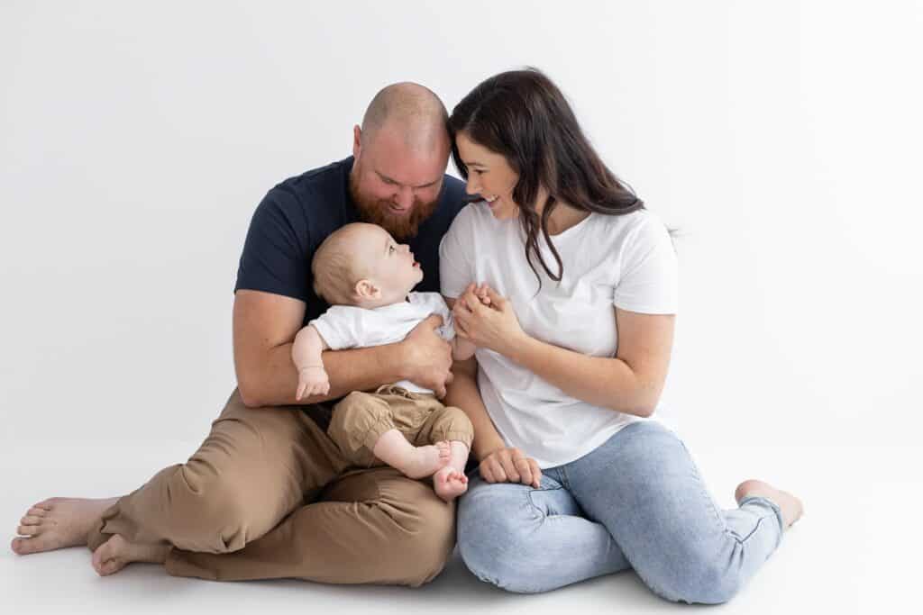 Parent sitting close together on the ground both looking down adoringly at their little boy during family photography session in white studio in Pakenham, Melbourne, Victoria