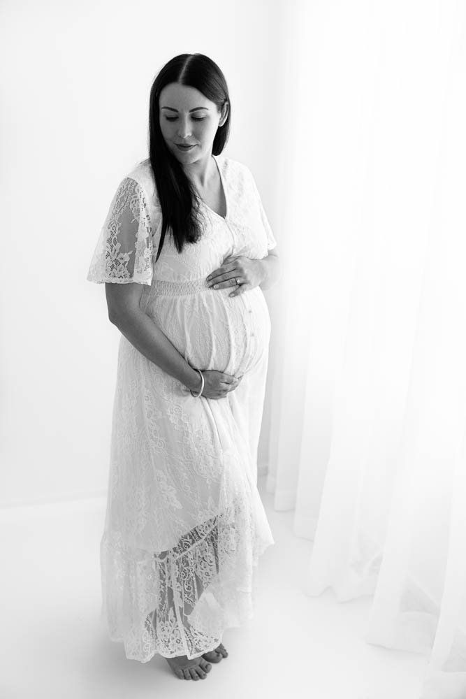 Pregnant mother in white lace dress cradles her belly while looking down in white photography studio in Melbourne