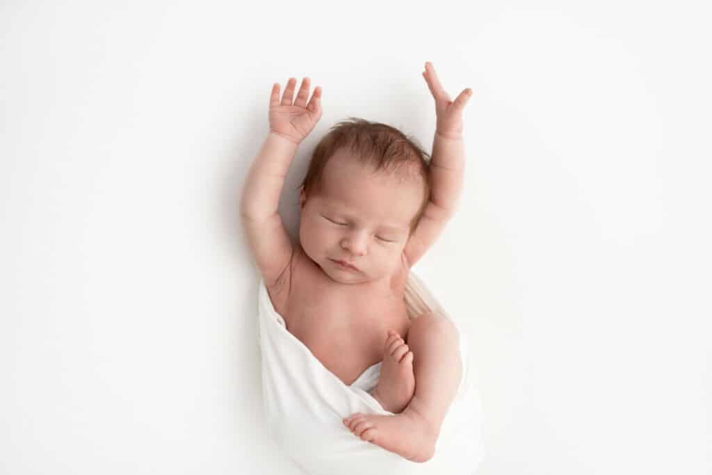 Newborn baby girl on white blanket stretching her arms up above her head during newborn photography session in Pakenham studio