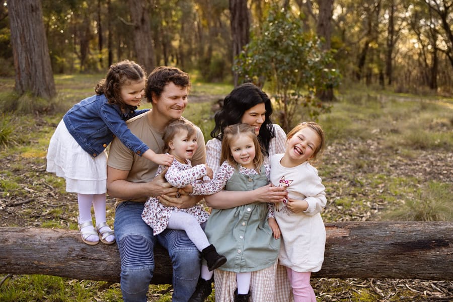 Family with Mum, Dad and four young girls sitting on a fallen tree all laughing while tickling one of the girls during outdoor sunset family photography session in Pakenham