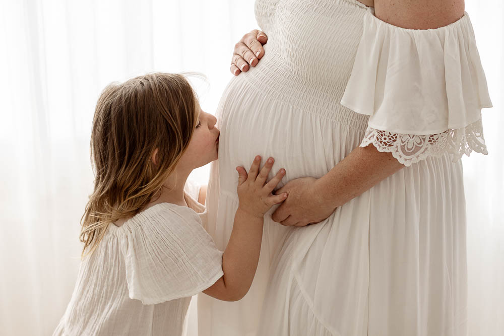 Daughter kissing and holding her Mothers pregnant belly, with both wearing white dresses in Melbourne Photography studio