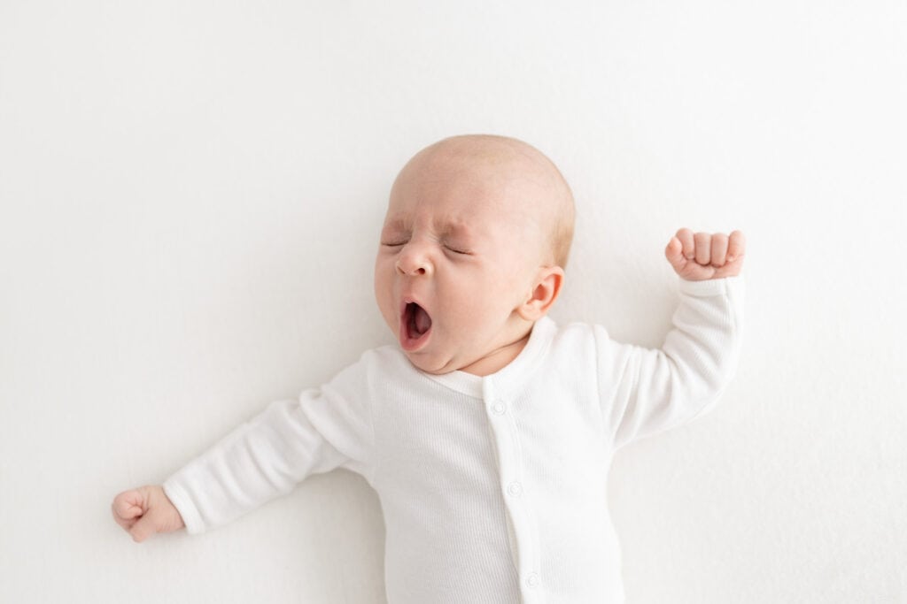 9 week old newborn baby in white button up onesie, stretching and yawning, while laying on white posing beanbag during newborn photoshoot in Melbourne