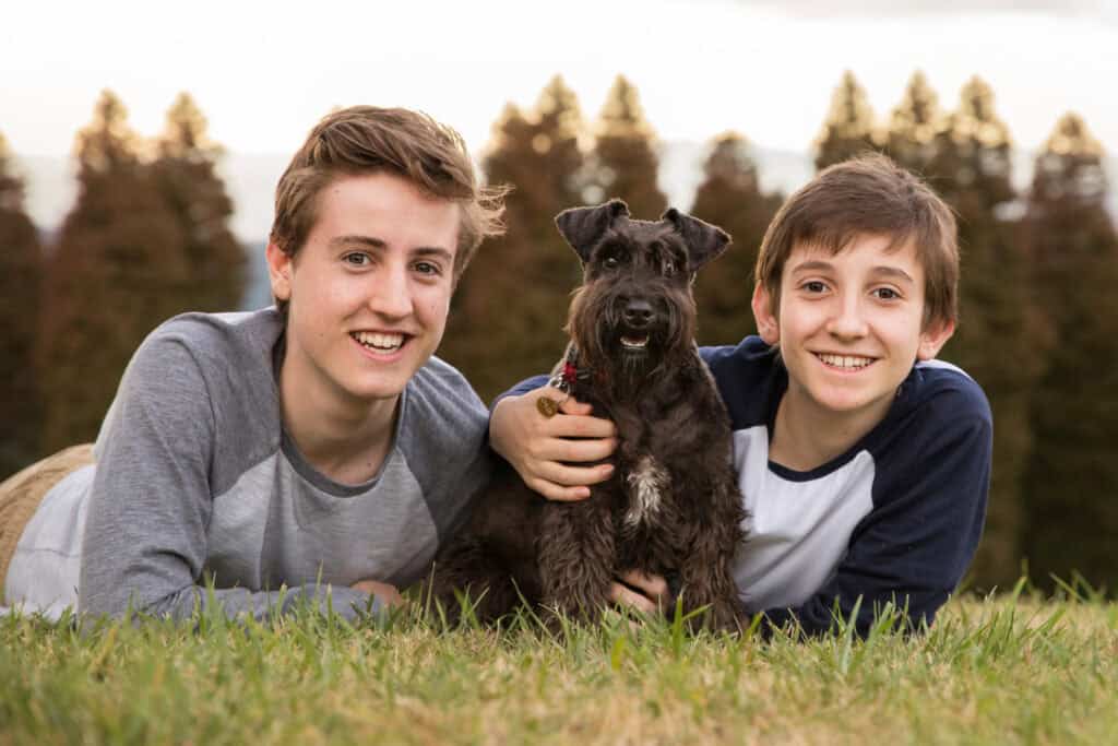 Black miniature schnauzer sitting with two human brothers laying on the ground with arms around her taken at Olinda in the Dandenong Ranges of Victoria