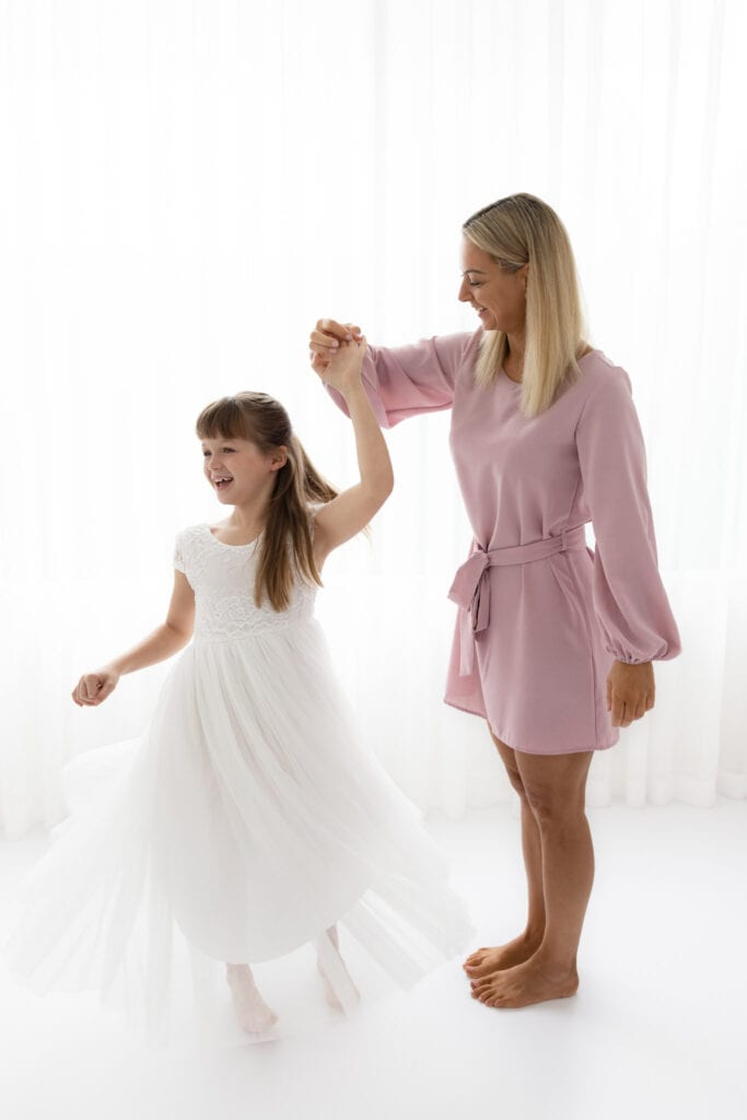 Mother in pink dress holds her daughters hand as she twirls in a long white dress in white melbourne photography studio